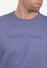 Hombre vistiendo un sudadera morada de cuello redondo con texto en relieve "AMARO JEANS" en el pecho, mirando ligeramente hacia la derecha.
