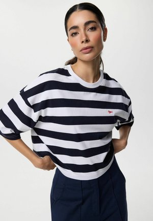 Kobieta z ciemnymi włosami związanymi z tyłu, ubrana w czarno-białą koszulę w paski i granatowe spodnie, stojąca z rękami na biodrach.