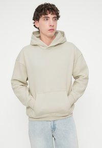 Φωτεινή μπεζ μπλούζα με κουκούλα από μαλακό ύφασμα, με μεγάλη μπροστινή τσέπη, ρυθμιζόμενη κουκούλα και άνετη εφαρμογή. Συνδυάζεται με ανοιχτόχρωμα μπλε τζιν.