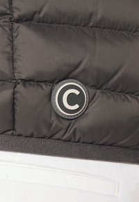 Svart quiltad tyg med en cirkulär lapp som har ett stort vitt bokstav "C" omgiven av svart, ovanför vitt texturerat tyg.