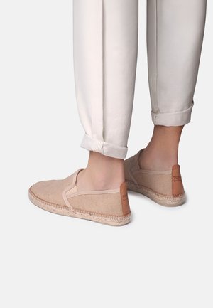 Pies con alpargatas de lona beige sin cordones combinadas con pantalones beige claro remangados sobre un fondo claro y liso.