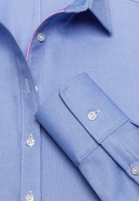 Helles blaues Hemd mit Knöpfen aus strukturiertem Stoff. Verfügt über weiße Knöpfe und einen kontrastierenden pinken Akzent im Kragen und an den Manschetten.