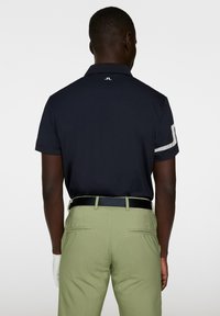 Navy polo shirt with short sleeves, featuring a white side accent; paired with light green trousers and a black belt. Golf glove on left hand.