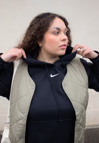 Prešívaná olivovo zelená vesta na čiernej mikine s kapucňou a bielym logom Nike, s možnosťou stiahnutia šnúrkou a rebrovanými manžetami; textúrovaná tkanina vytvára kontrast.