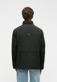 Mustat vedenpitävät takki, jossa on korkea kaulus ja takana logo, jossa on punaisia, valkoisia ja sinisiä yksityiskohtia. Kevyt kangas ja rento malli.