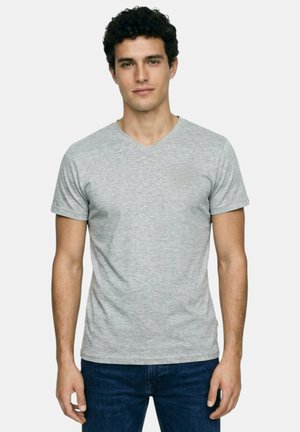 Jeune homme aux cheveux bouclés et foncés, portant un t-shirt gris clair uni à col en V et un jean bleu foncé, debout devant un fond neutre.