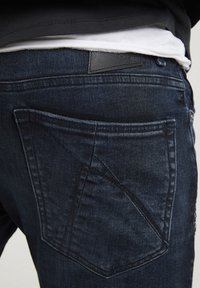 Detailní záběr na osobu v tmavě modrých džínech s zadní kapsou a viditelným švem, doplněných bílou košilí a černým topem.