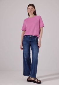 Haut rose à manches courtes, coupe décontractée, associé à un jean bleu à jambes larges. La mannequin porte des sandales noires avec des accents métalliques. Apparence lisse et décontractée.