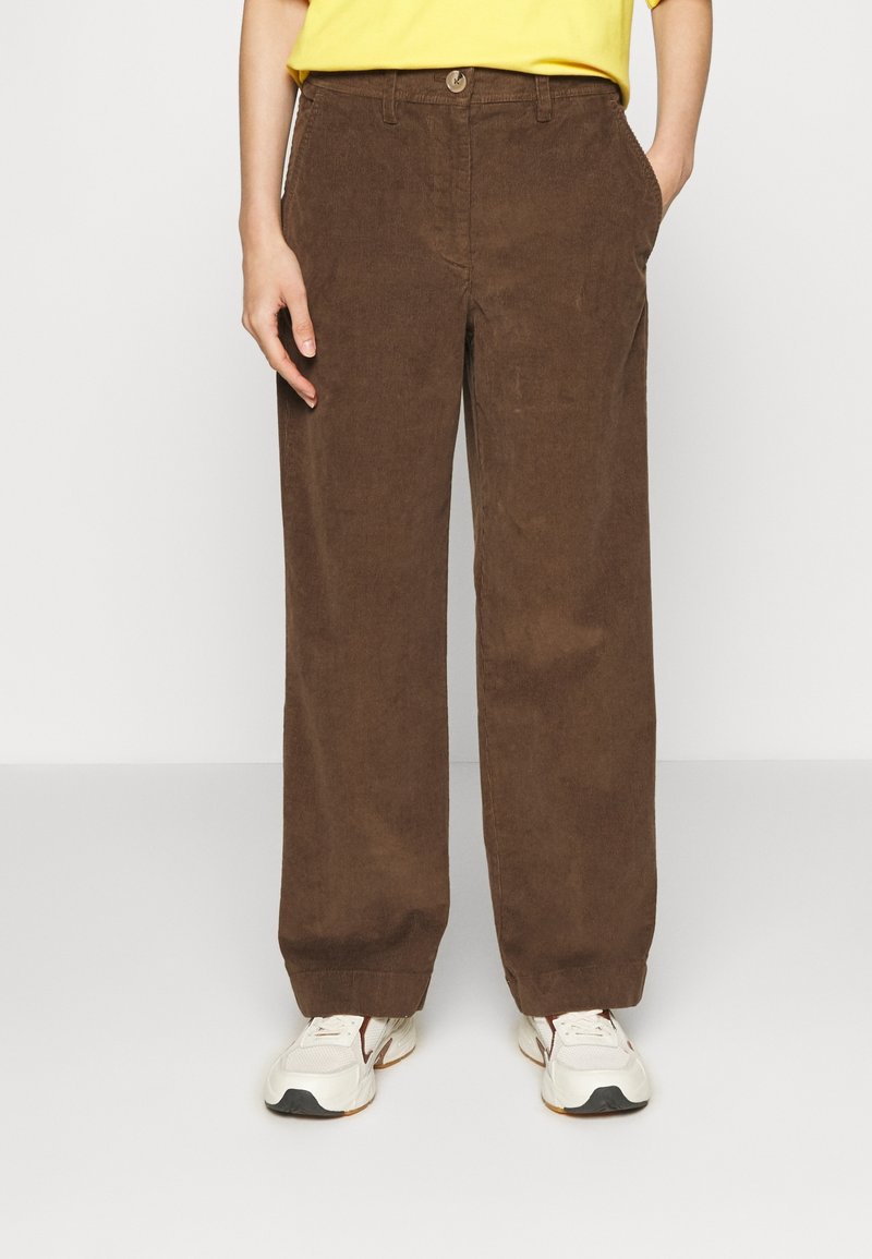 Personne portant un pantalon large en velours côtelé marron, un haut jaune et des baskets blanches, se tenant contre un fond blanc uni.