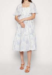 Biała sukienka w kwiaty z niebieskimi wzorami, krótki falbaniasty rękaw, kwadratowy dekolt i warstwowa spódnica. W zestawie z pomarańczowymi sandałami na paski.