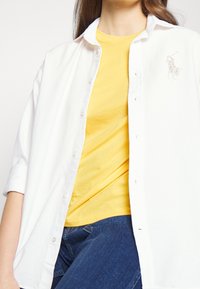 Vit skjorta med uppvikta ärmar över en gul t-shirt. Har en liten, glittrande logotyp på vänster bröst. Denimblå jeans synliga.