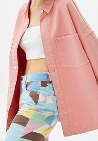 Rosa denimskjorta med tryckknappar, oversized passform, bärs över en vit ribbad crop top. Mönstrade blå jeans med geometriska former i pastellfärger.