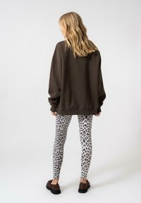 Sötétbarna, oversized kapucnis felső, szürke leggings-szel, amely fekete leopárdmintával rendelkezik. A modell háttal áll a kamerának.