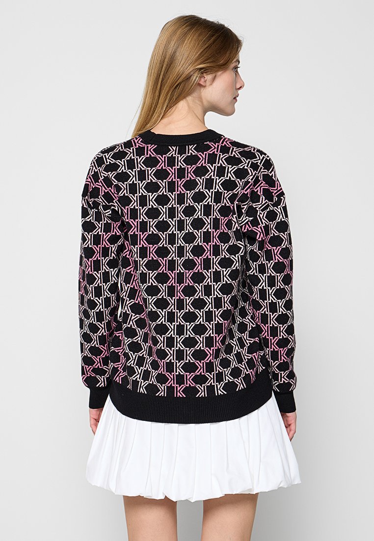 Schwarzer, gestrickter Pullover mit einem geometrischen Muster in Rosa und Weiß. Hat einen runden Halsausschnitt und gerippte Ärmelbündchen. Trägt dazu einen weißen, plissierten Rock.