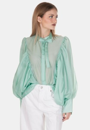 Donna con una camicetta verde menta translucida con maniche lunghe a sbuffo e colletto con fiocco, abbinata a pantaloni bianchi a vita alta, che guarda di lato.