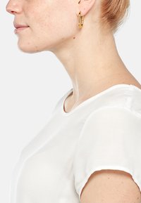 Orecchino a cerchio in oro dal design minimalista, realizzato in metallo lucido. Il modello indossa una maglietta bianca a maniche corte con scollatura rotonda.