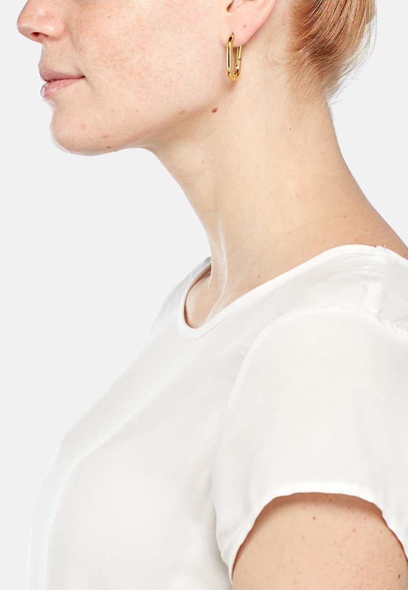 Orecchino a cerchio in oro dal design minimalista, realizzato in metallo lucido. Il modello indossa una maglietta bianca a maniche corte con scollatura rotonda.