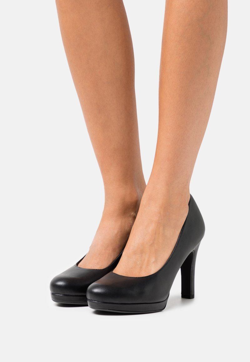 Jambes portant des talons hauts compensés en cuir noir avec bouts arrondis sur un fond clair.