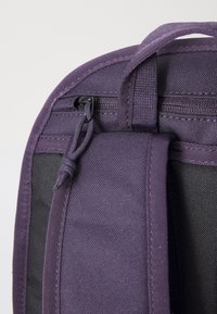 Canto de uma mochila de tecido roxo mostrando zíperes, costura e um detalhe em laço; apresenta um acabamento texturizado com um lado preto contrastante.