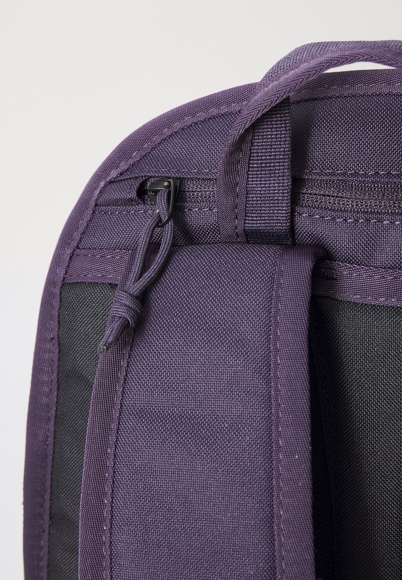 Un coin d'un sac à dos en tissu violet montrant les zips, les coutures et un détail en boucle; présente une finition texturée avec un côté noir contrastant.