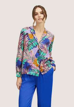 Camicia a maniche lunghe con un colorato motivo di foglie tropicali su sfondo scuro; presenta un colletto e una chiusura con bottoni sul davanti. Abbinata a pantaloni blu.