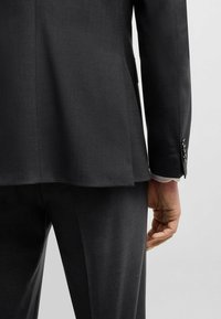 Dunkelgrauer Blazer und Hose aus strukturiertem Stoff. Der Blazer hat einen einfachen Schlitz, und die Ärmel verfügen über Knopfakzente.