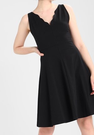 Nainen pukeutuneena hihattomaan mustaan mekkoon, jossa on aaltoileva V-pääntie, poseeraa yhdellä kädellä lantiolla yksinkertaista taustaa vasten.