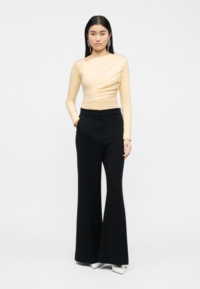 Γυναίκα με μακριά μαύρα μαλλιά φοράει ασύμμετρο ανοιχτό μπεζ τοπ, μαύρο παντελόνι φαρδύ, λευκά μυτερά παπούτσια και κρεμαστά σκουλαρίκια, όρθια.