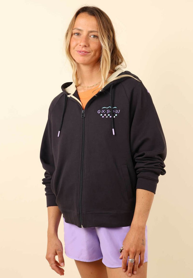 Haut à zip noir avec doublure crème, présentant un logo imprimé violet et vert sur la poitrine. Manches longues, poches kangourou, coupe décontractée.