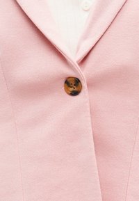 Blazer rose pâle fermé par un seul bouton en écaille de tortue marron, laissant apercevoir une partie d'une chemise blanche côtelée en dessous.