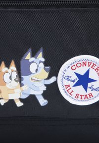 Juodas tekstilinis maišelis su animacinių šunų iliustracijomis ir baltu apvaliu lipduku su raudonu tekstu ir mėlynu žvaigžde, pažymėtu „Converse All Star“. 