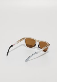 Ochelari de soare cu un cadru transparent de culoare marron, lentile marron, brațe albe și negre, având un logo pe o braț și accente din cauciuc pentru aderență.