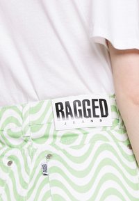 Jeans verdes e brancos com um padrão ondulado. Apresentam uma etiqueta preta proeminente com a inscrição "RAGGED JEANS". Botões de metal visíveis.