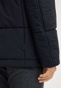 Giubbotto imbottito blu navy con motivo a trapunta; presenta un'etichetta con il logo sul maniche; indossato sopra pantaloni grigi con motivo.