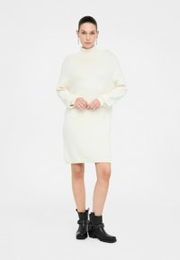 Abito bianco lavorato a maglia con collo alto e maniche lunghe, abbinato a stivali neri alla caviglia con hardware borchiato e tacco basso.