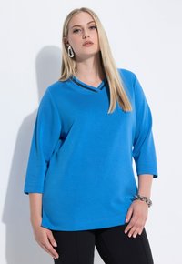 Camiseta azul con escote en V y mangas tres cuartos, con un cuello de contraste. Textura suave, corte casual y aberturas laterales.