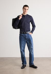 Navy button-up shirt, light blue jeans, black belt, and black shoes. Model carries a textured black tote bag. Neutral background.