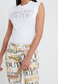 Bílý bezrukávový tričko s kamínkovým nápisem "VERSACE JEANS COUTURE". V kombinaci s vzorovanými, roztrhanými džíny s zlatými akcenty.