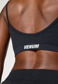 Crni sportski grudnjak od rastezljivog materijala, s širokim pojasom na kojem je u bijelom ispisano "VENUM". Glatka tekstura i minimalistički dizajn.