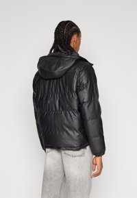 Czarna puchowa kurtka z kapturem, z pikowanym wzorem, błyszczącym wykończeniem, elastycznymi mankietami i luźnym krojem. Noszona na jasnoszarych spodniach.