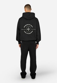Μαύρη hoodie με κουκούλα και κορδόνι, με εκτυπωμένο σχέδιο πυξίδας στην πλάτη. Συνδυασμένη με μαύρες υφασμάτινες joggers που ταιριάζουν.