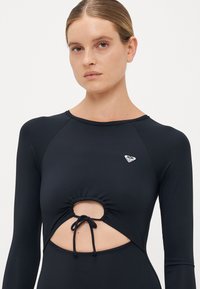Woman wearing a black long-sleeve top with a central cutout and tie detail, featuring a small white logo on the chest.