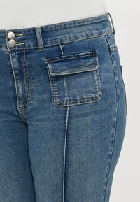 Denim bikses ar izbalējušu zilu krāsu, divu pogu aizdari un vienu kabatu ar vāku aizmugurē. Tekstūra ir gluda ar viegli nodiluma rakstiem.