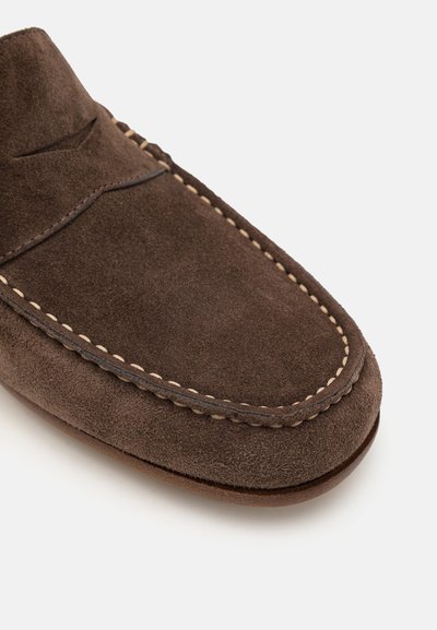Mocassin en suède marron avec des détails de couture visibles et un bout arrondi, présentant une surface texturée et un design bas.