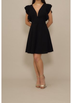 Femme portant une robe noire trapèze avec des manches volantées et des talons à brides dorées, debout devant un fond beige uni.