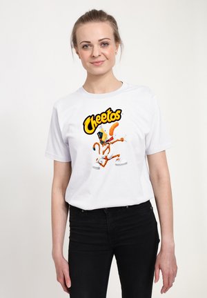 Młoda kobieta z włosami związanymi do tyłu, ubrana w białą koszulkę Cheetos i czarne spodnie, stoi na tle jednolitego jasnego tła.