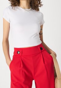 Biała dopasowana crop top połączona z czerwonymi spodniami o wysokim stanie, wyposażonymi w boczne kieszenie i czarne guziki. Gładka tekstura materiału.