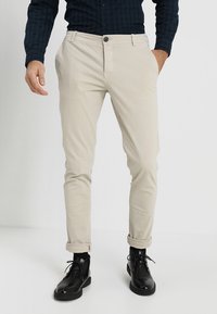Beige chinos med slim fit, knappstängning och uppvikta fållar. Bärs med svarta snörkängor och en mörk rutig skjorta.