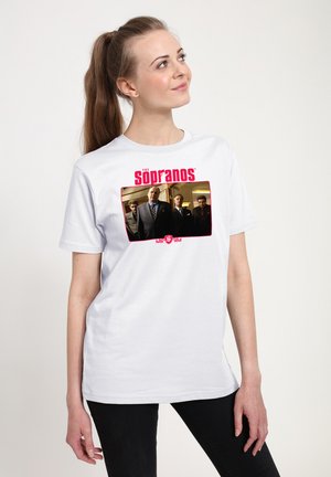 Kvinde i hvid T-shirt med Sopranos-castet, står indendørs, ser op og smiler, iført sorte bukser og hestehale.