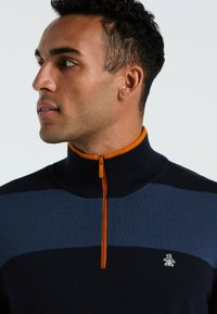 Marineblauwe gebreide pullover met een oranje rits en kraag. Heeft horizontale strepen in donkerblauw en lichtblauw, met een pinguïnlogo op de borst.